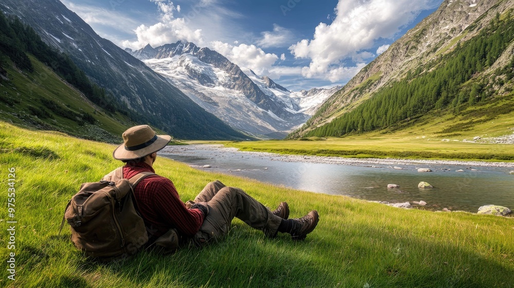 Naklejka premium A hiker sits on a grassy meadow beside a river with snow-capped mountains in the distance.