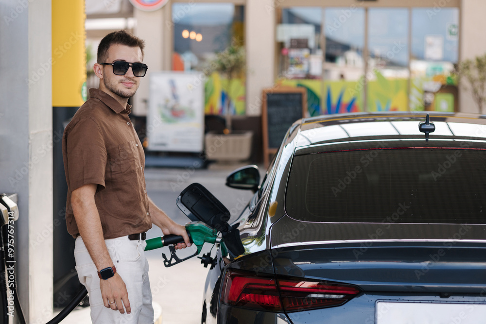 Fototapeta premium Handsome man in sunglasses refuelling his car on self service gas station. Male filling diesel at gasoline fuel and look into camera