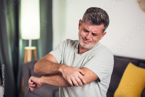 Man With Pain In Elbow. Pain relief concept. Middle age man feeling the pain in hand elbow at home. Male touching his painful elbow photo