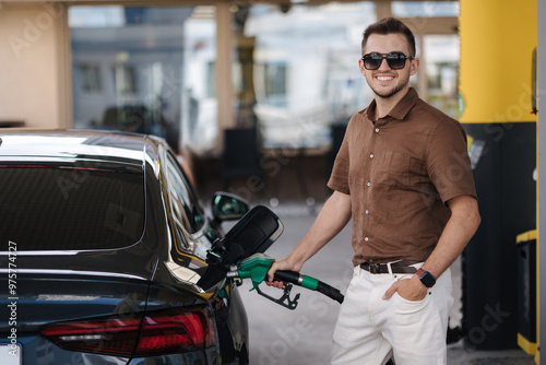 Smiling man refuelling his luxury car at the gas station. Guy looking camera and pouring petrol into tank of modern vehicle on filling station in city