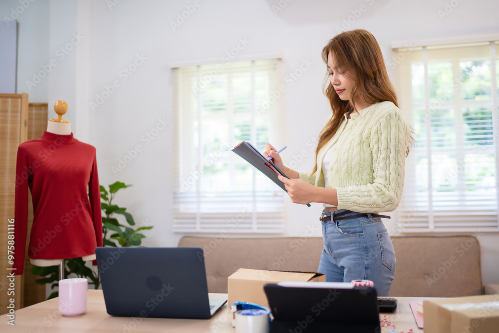 A young woman running an online clothing business is standing at her desk, holding a tablet and reviewing orders. She is surrounded by boxes, laptops, and clothing items.