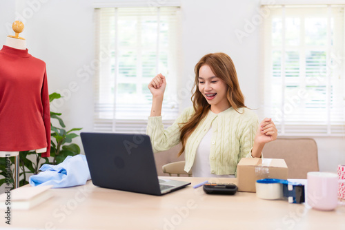 A young woman managing her online clothing business is excited as she works on her laptop. She is surrounded by clothing, packaging materials, and is in a bright, organized workspace