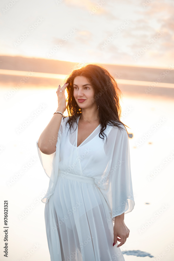  girl with black curly hair in a white dress on the shore of the lake in the sunset light. calm sensual portrait. 