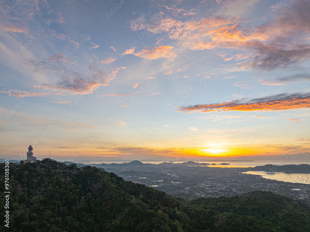 Fototapeta premium Arial view stunning sunrise above Phuket big Buddha on the mountain. A breathtaking panoramic view of a vibrant sunrise casting warm hues over a coastal town, with the sea reflecting of the sky