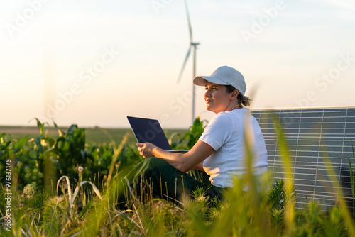 Woman farmer wearing white cap and t-shirt with laptop sits next to solar panel at sunset. Wind turbines in the background