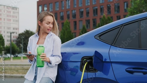 Young woman with refillable water bottle and cellphone charges EV at city station. Female electric car owner shows modern technology and sustainable living