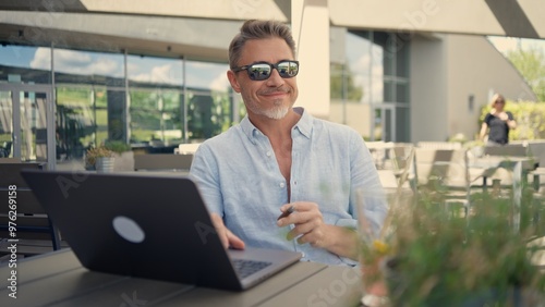 Mature male freelancer working on laptop computer outdoor. Happy man sitting at cafe, smiling. Businessman enjoying freedom in work and connectivity in age of Internet.