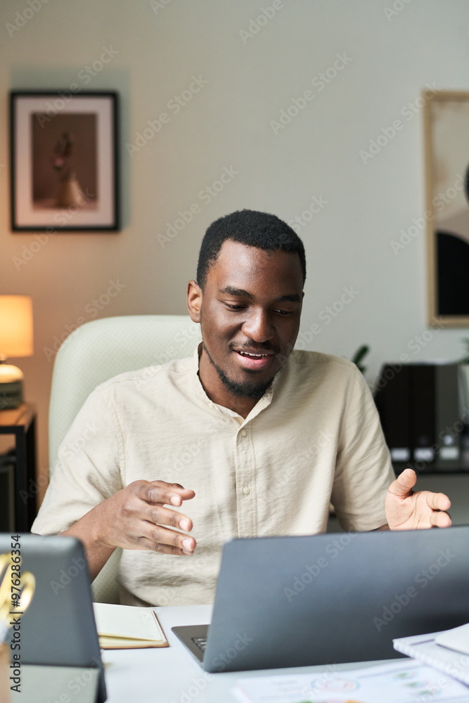 Smiling man sitting at desk in office setting, using laptop. Engaging in conversation while holding gesturing hand in relaxed office environment Smiling man sitting at desk in office setting, using laptop. Engaging in conversation while holding gesturing hand in relaxed office environment
