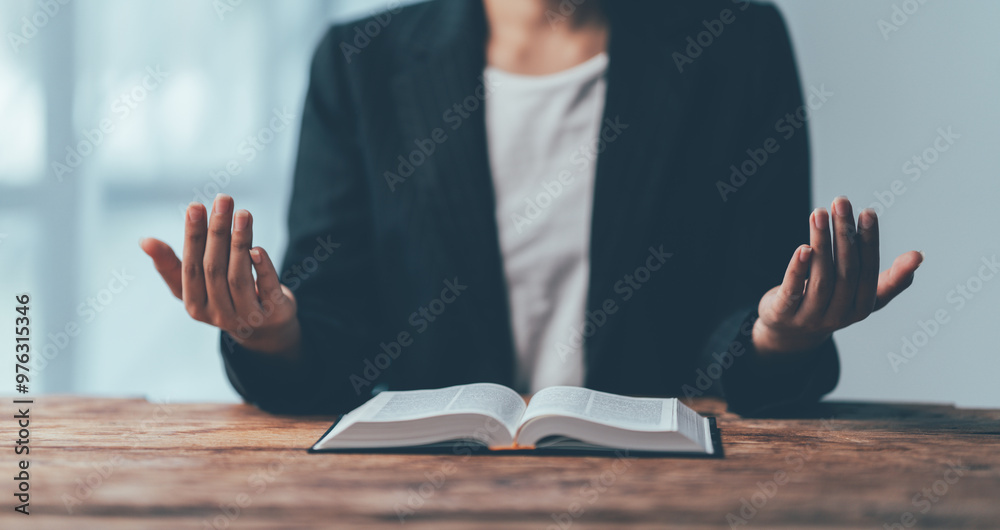Obraz premium Open Hands, Open Heart: A woman in a business suit sits at a desk with an open book, her hands raised in a gesture of prayer, symbolizing faith, hope, and devotion. The light streaming in from the win