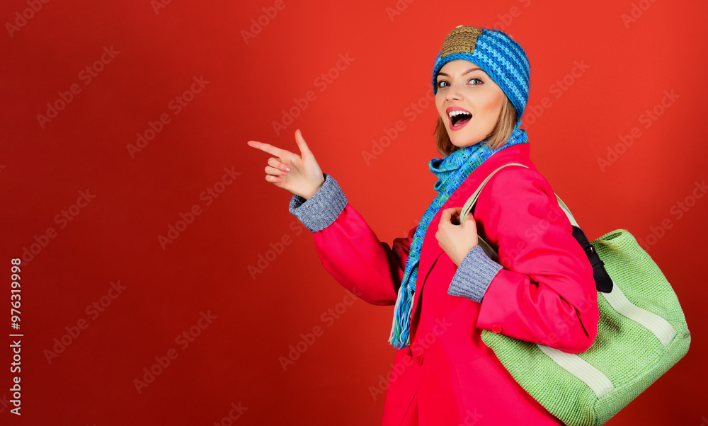 Smiling woman in blue hat, scarf and red coat with green knitted handbag pointing finger at copy space. Fashion spring autumn winter. Stylish girl in fashionable clothing. Season sales and discounts. Smiling woman in blue hat, scarf and red coat with green knitted handbag pointing finger at copy space. Fashion spring autumn winter. Stylish girl in fashionable clothing. Season sales and discounts.