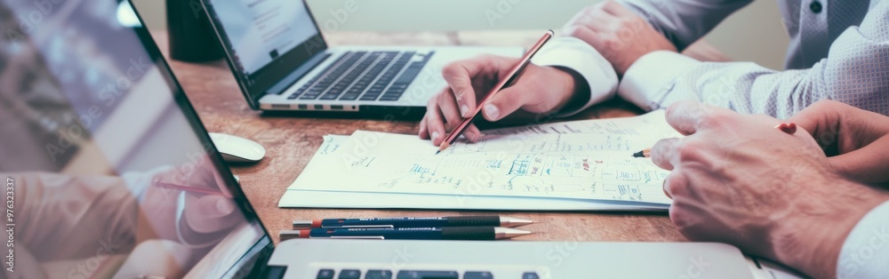 Two people working together at a desk with laptops, documents, and pencils, collaborating on a project in an office setting Two people working together at a desk with laptops, documents, and pencils, collaborating on a project in an office setting