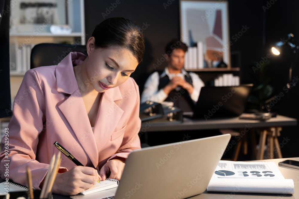 Focusing on sitting woman coworker over thinking campaign writing on paperwork and laptop on working desk while man leaning at desk waiting email at late night time with blurry background. Postulate. Focusing on sitting woman coworker over thinking campaign writing on paperwork and laptop on working desk while man leaning at desk waiting email at late night time with blurry background. Postulate.