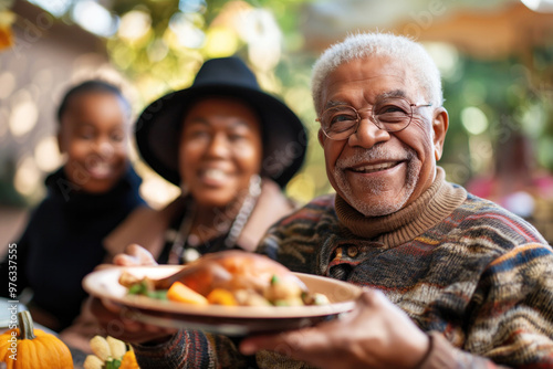 Elderly black man holding a plate with roasted turkey, with his family blurred in the background, everyone smiling and enjoying the festive atmosphere outdoors.