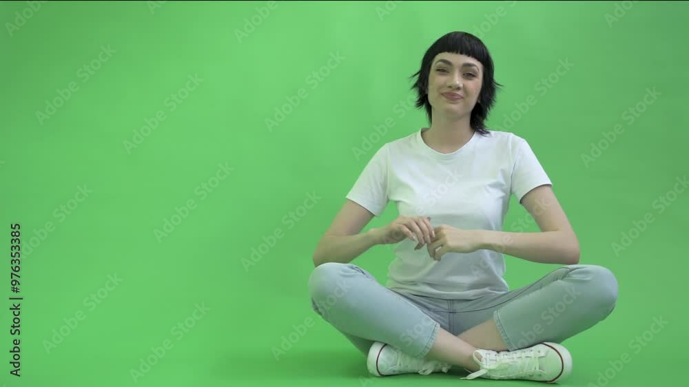 Young Woman Sitting Сross-legged in Jeans Pointing to Empty Place on Green Background Young Woman Sitting Сross-legged in Jeans Pointing to Empty Place on Green Background