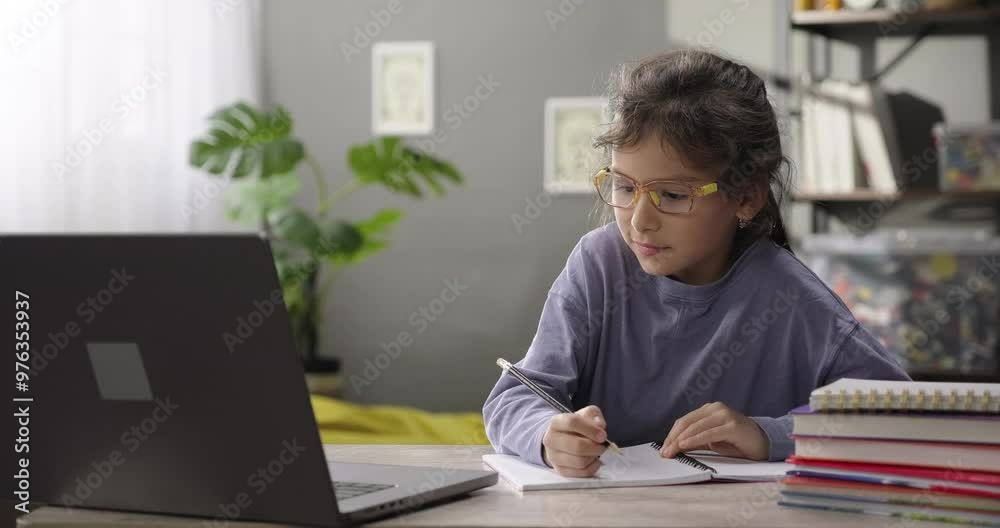 Cute little girl e-learning online using app or website on digital tablet. Focused girl looking at the screen, watching educational tutorial, making notes in a copybook sitting at the desk at home Cute little girl e-learning online using app or website on digital tablet. Focused girl looking at the screen, watching educational tutorial, making notes in a copybook sitting at the desk at home