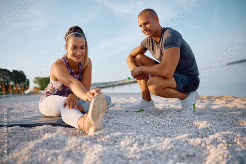 Happy sportswoman stretching her leg while exercising with  friend on beach. photo