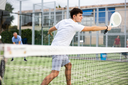 Positive focused guy playing friendly paddleball match on outdoor court on summer day, confidently hitting ball with backhand. Young people sports concept photo