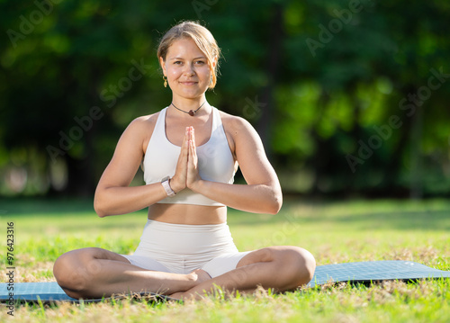 Girl in park on green grass of lawn performs asanas of hatha yoga exercises. She practicing lotus position, padmasana. Concept of relaxation and moderate physical activity during walks photo