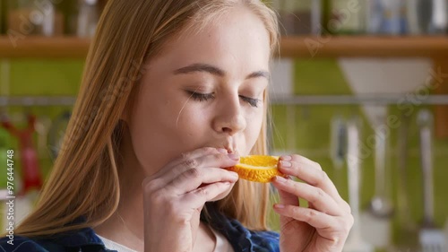 Woman eating juicy slice of orange at kitchen, close up portrait