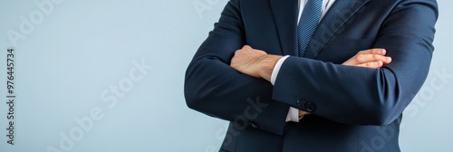 A man dressed in a formal business suit is standing with his arms crossed against a neutral background, exuding confidence and professionalism.