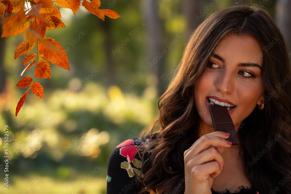 Fototapeta premium Closeup shot of amazing beautiful brunette biting dark chocolate in the park, fall season