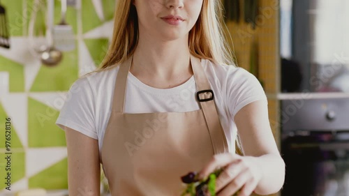 Young woman preparing fresh salad at home, adding cut greenery to bowl