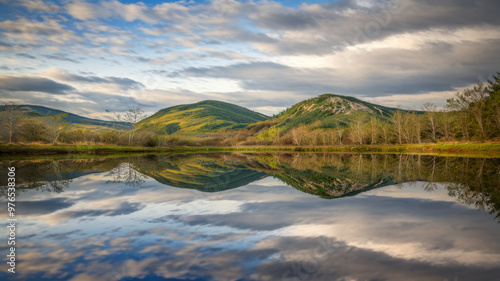 A photo of a serene landscape with a calm lake reflecting the sky, mountains, and trees. The sky is a mix of blue and grey clouds. The mountains have green trees and the ground is covered with grass. 