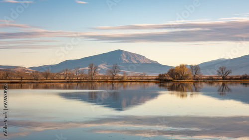 A photo of a vast, serene landscape with a calm lake reflecting the sky and trees. There are mountains in the background with a few trees near the lake. The sky is clear with a few clouds.