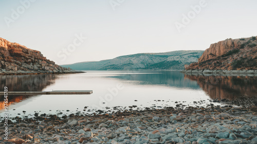 A photo of a serene landscape with a vast body of water, surrounded by rocky cliffs and a clear sky. The water is calm, reflecting the sky and the cliffs. There is a wooden dock