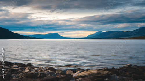 A photo of a serene landscape with a vast body of water leading to the horizon. The sky is overcast with dark clouds. There are mountains in the background. The foreground contains rocks.