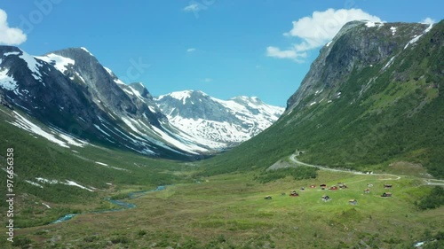 Aerial view of a vast green valley surrounded by towering mountains in Norway and cabin community in lowlands, with patches of snow on the peaks under a clear blue sky. photo