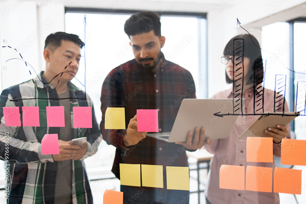 © sofiko14 - Team members collaborating in brainstorming session with sticky notes on glass wall analyzing charts and data diagrams. Clear focus on teamwork and productivity in modern office environment.