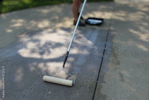 a paint roller for sealing being used on a concrete driveway with a persons legs and paint tray in the background