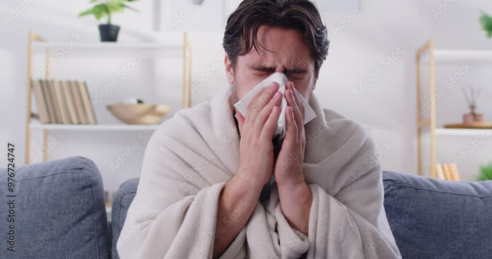 Unwell man wrapped in a blanket, sneezing and suffering from cold or flu symptoms. He is sick, with a fever, measuring his temperature with a thermometer as he battles a viral disease. Unwell man wrapped in a blanket, sneezing and suffering from cold or flu symptoms. He is sick, with a fever, measuring his temperature with a thermometer as he battles a viral disease.