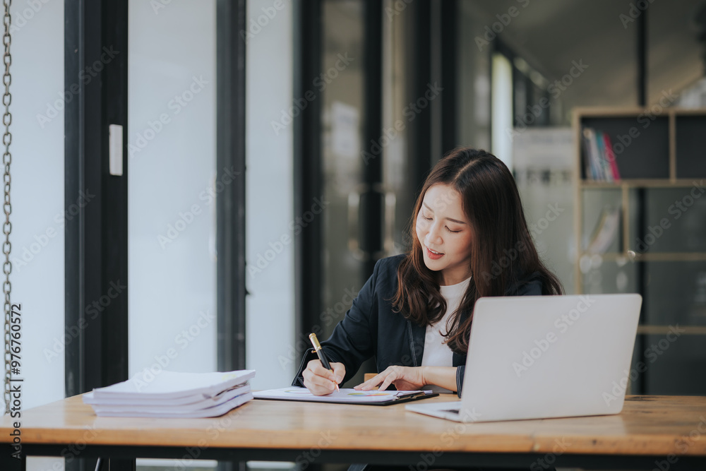 A cheerful young Asian businesswoman sits at her modern desk, typing on a laptop, working on corporate projects with paperwork, smiling in a busy workplace. A cheerful young Asian businesswoman sits at her modern desk, typing on a laptop, working on corporate projects with paperwork, smiling in a busy workplace.