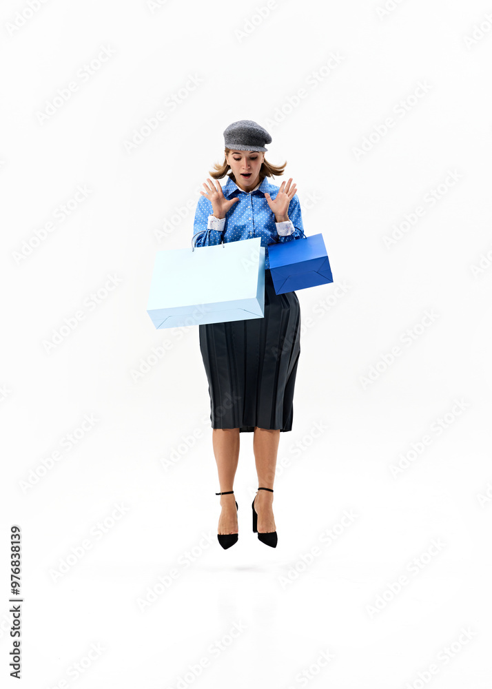 Woman looks surprised as she jumps holding two shopping bags, both hands raised with excitement, posing in chic outfit against white studio background. Concept of shopping, Black Friday, fashion. Ad Woman looks surprised as she jumps holding two shopping bags, both hands raised with excitement, posing in chic outfit against white studio background. Concept of shopping, Black Friday, fashion. Ad
