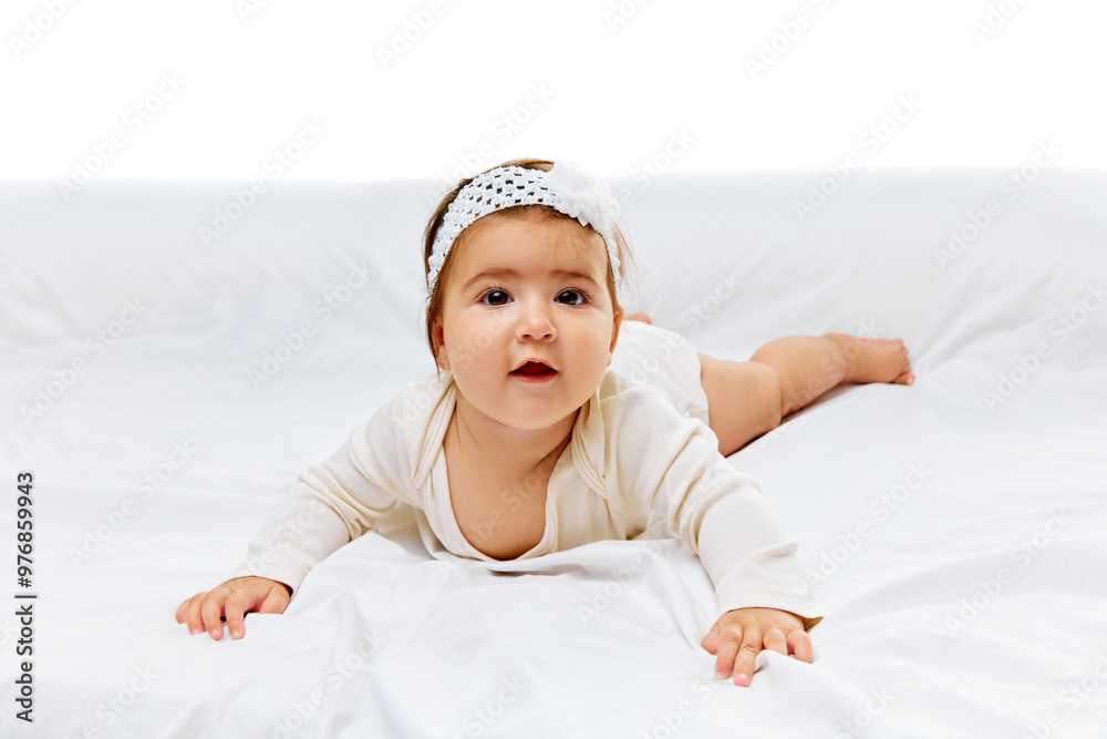 Sweet girl in white onesie and floral headband gazes up, resting comfortably on soft bed against white studio background. Concept of beauty, parenthood, childhood, life and birth. Sweet girl in white onesie and floral headband gazes up, resting comfortably on soft bed against white studio background. Concept of beauty, parenthood, childhood, life and birth.