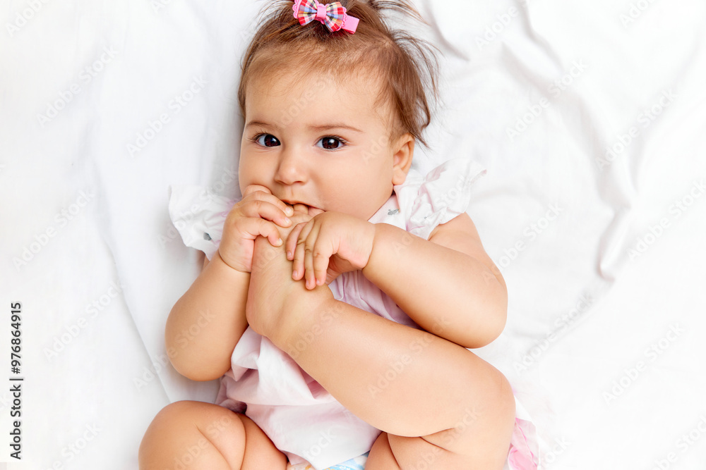 With playful smile, baby girl puts her foot in her mouth, showing her adorable innocence while lying comfortably on bed. Concept of beauty, parenthood, childhood, life and birth. With playful smile, baby girl puts her foot in her mouth, showing her adorable innocence while lying comfortably on bed. Concept of beauty, parenthood, childhood, life and birth.