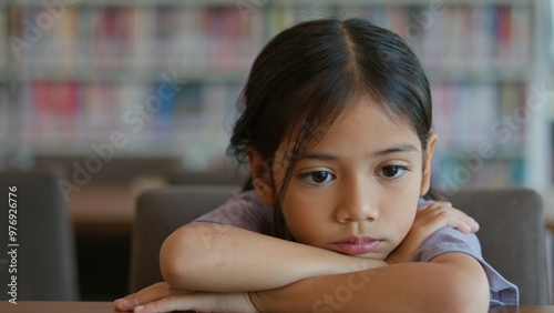 An Asian girl sits at a table in a library. She looks bored and lost in thought. Her disinterested expression shows her disinterest in the things around her. photo