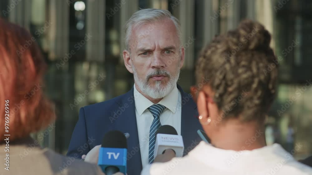 Waist up shot of confident mature Caucasian male politician wearing blue suit, white shirt and blue striped tie giving interview to TV reporters while standing outdoors Waist up shot of confident mature Caucasian male politician wearing blue suit, white shirt and blue striped tie giving interview to TV reporters while standing outdoors
