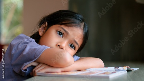 An Asian girl sits at a table in a library, her face clearly showing boredom as she looks away from the open book in front of her. She looks disinterested and lost in thought.