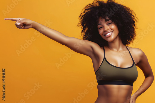 Vibrant fitness portrait of a smiling woman with curly hair, pointing confidently to the side in a sports bra against a bright orange background, symbolizing energy, motivation, and a healthy lifestyl photo