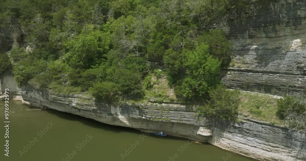 Person in a blue kayak paddles beside a rocky cliff, exploring a calm waterway under shade Person in a blue kayak paddles beside a rocky cliff, exploring a calm waterway under shade