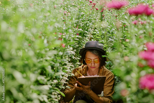 Person with glasses wearing hat immersed in reading notes in lush flower garden holding tablet. Pink flowers prominently frame lush greenery creating tranquil environment