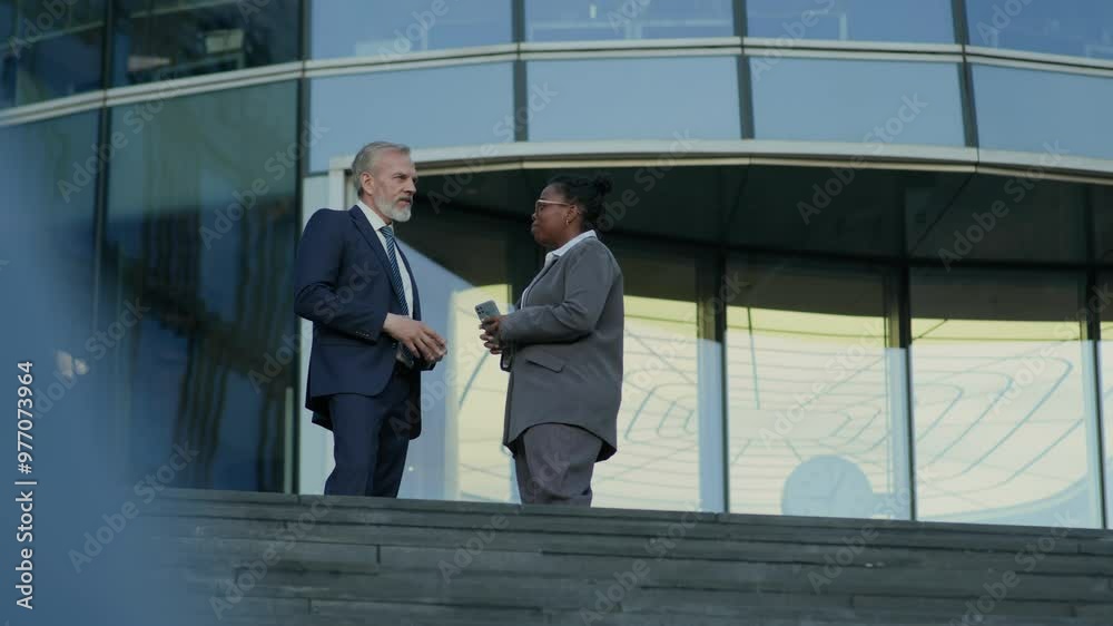 Low angle shot of successful ethnically diverse male and female business partners in formalwear standing on stairs outside modern building and having conversation Low angle shot of successful ethnically diverse male and female business partners in formalwear standing on stairs outside modern building and having conversation