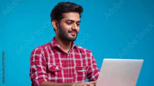 blue studio background, a young Indian IT specialist in a red shirt works on his laptop.