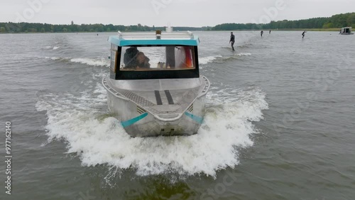 hydro foilers surfing wave behind watercraft driven by blond girl filmed with drone from front close up with trees on riverbank photo