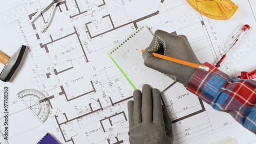 Close-up of a worker's hands writing on a notepad with a pencil, tools and a large blueprint lying nearby