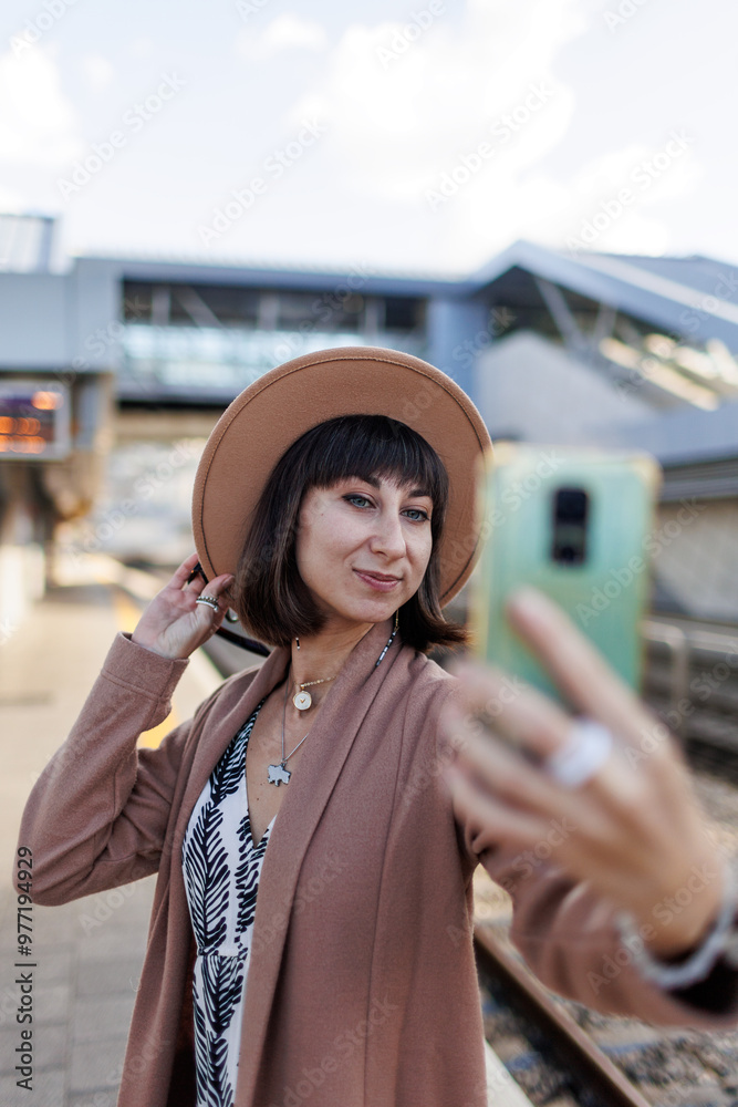Fototapeta premium girl waits for a train on the platform and takes a selfie. beautiful tourist woman in a hat takes a selfie at the train station. travel and adventure.