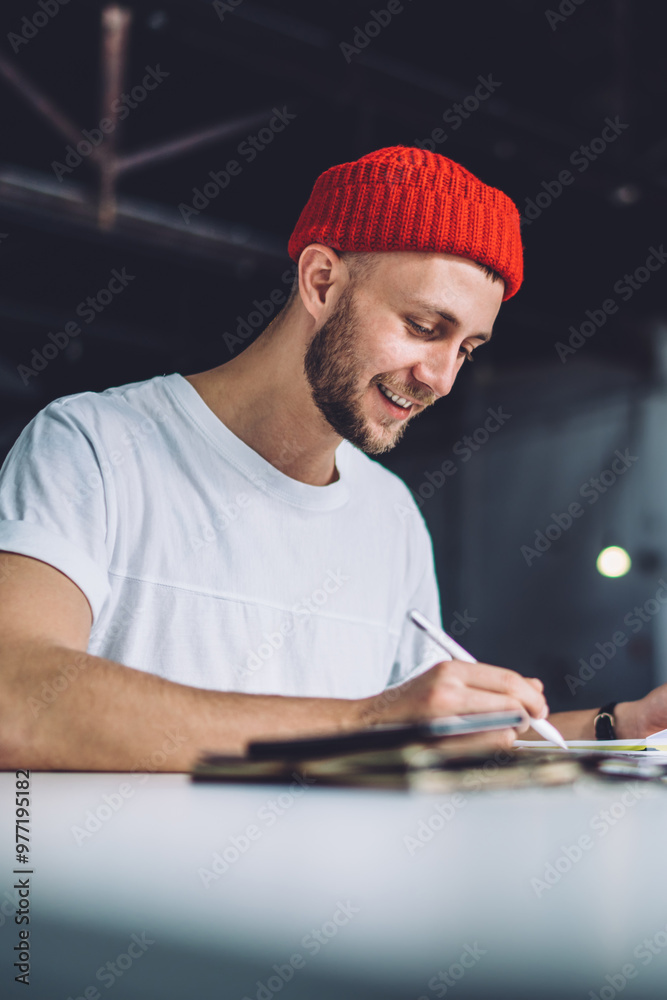 Handsome Caucasian man in trendy red cap making memo notes during learning process for preparing to college exams, positive hipster guy creating smm text for publication during working time Handsome Caucasian man in trendy red cap making memo notes during learning process for preparing to college exams, positive hipster guy creating smm text for publication during working time